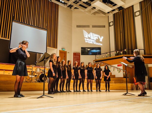 Choir of 10 girls and flautist being conducted by female condutor.